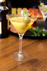 Chili-rimmed martini glass with yellow cocktail and olives.